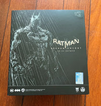 A boxed Batman Arkham Knight 1/12 scale figure package lying on a wooden floor. The box is predominantly black with a white sketch-style Batman figure and rain graphic, plus visible Batman and DC branding.