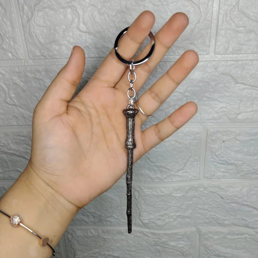 Hand holding a keychain shaped like a wand against a textured gray wall.