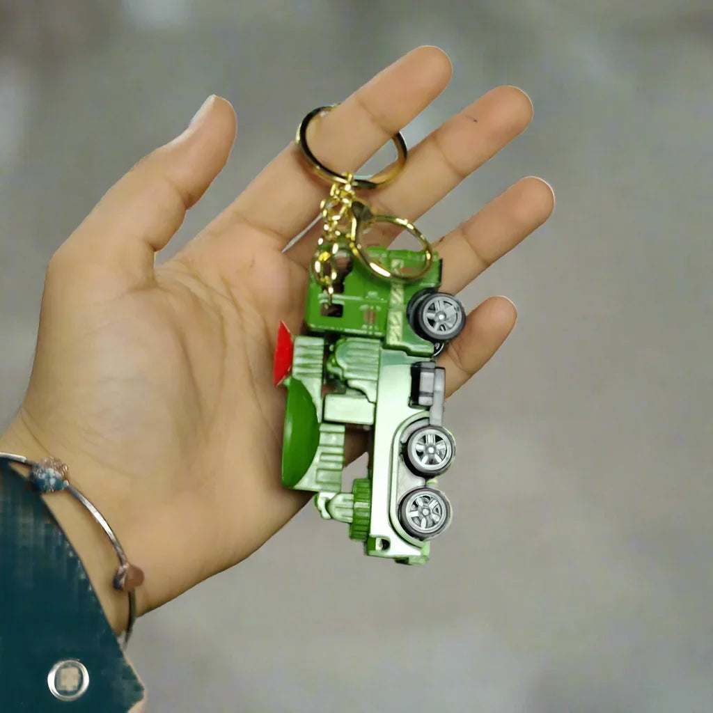 Hand holding a keychain with a green car design against a textured wall and grassy floor.