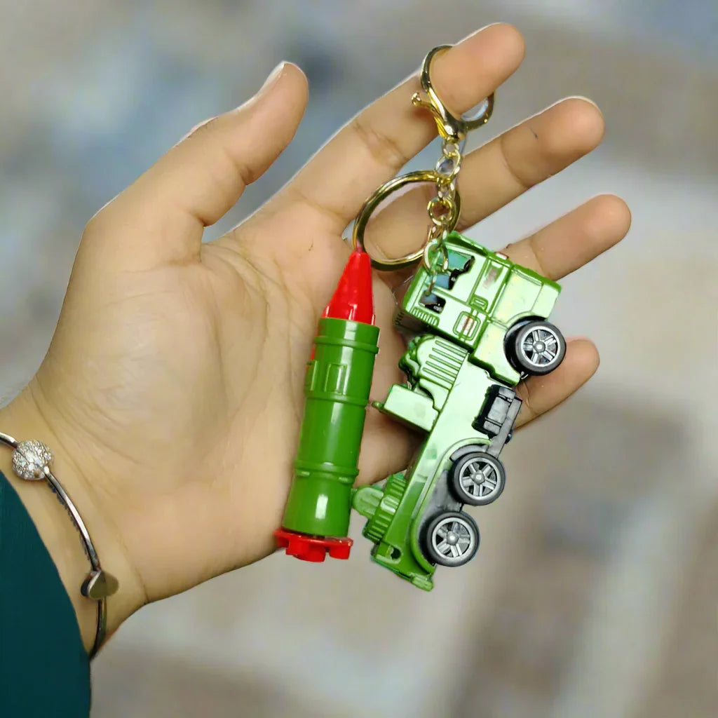 Hand holding a green toy truck keychain with red accents on a grassy surface.