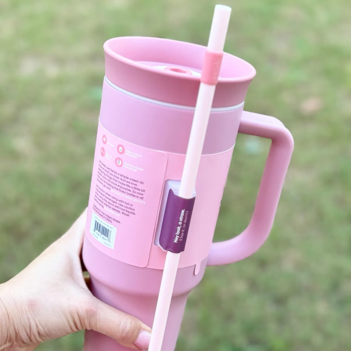 Pink insulated mug with handle and straw held by a hand against a blurred green background