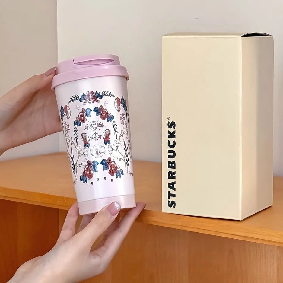 Starbucks mug with floral design held by a hand next to its packaging on a wooden surface.