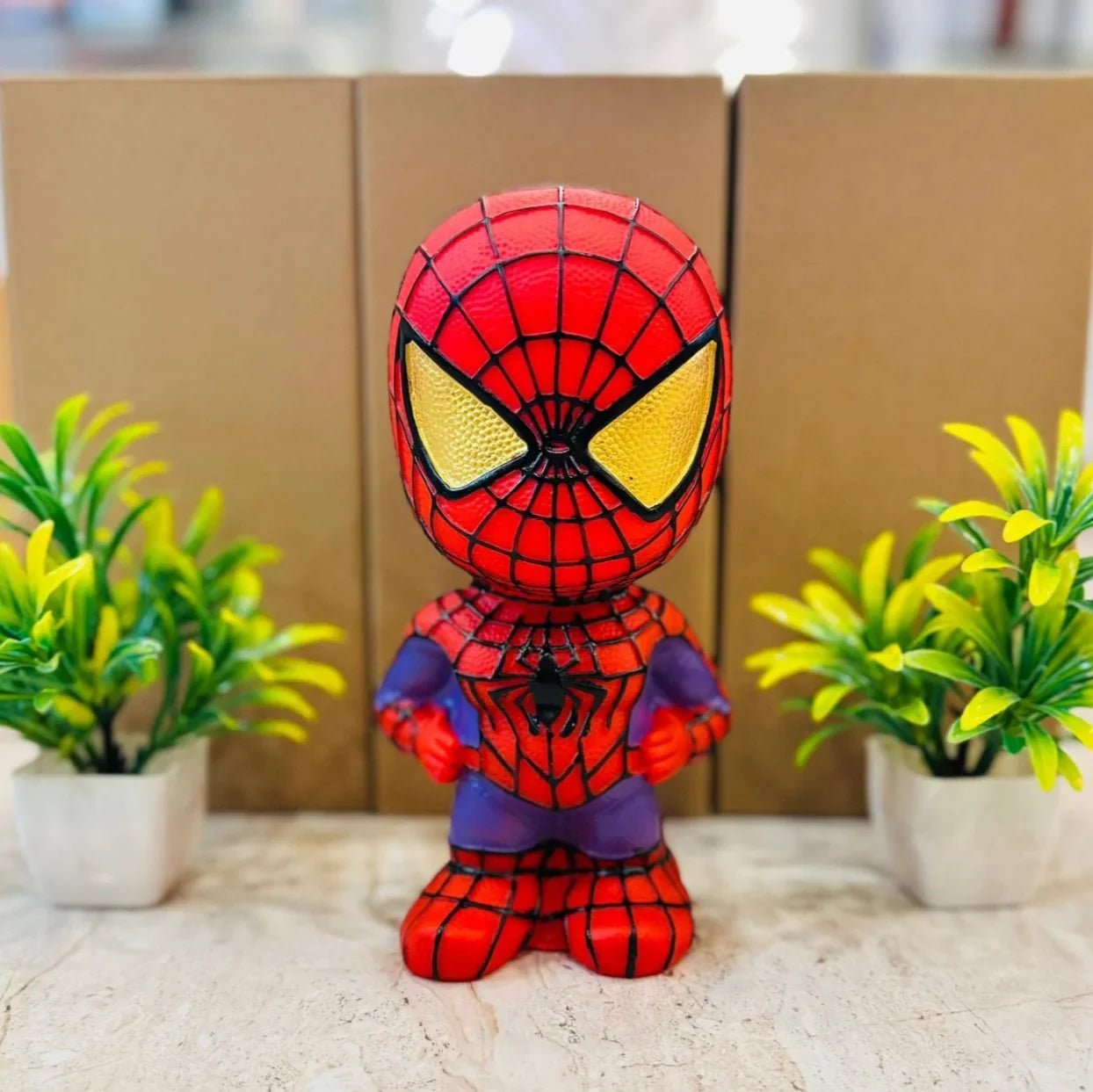 Spider-Man money bank figurine between two potted plants with cardboard boxes in the background