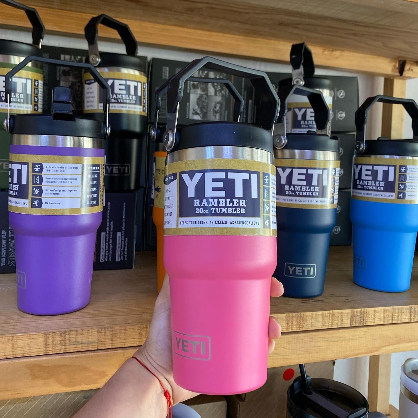 Hand holding a pink YETI rambler tumbler with more tumblers in the background on a wooden shelf.