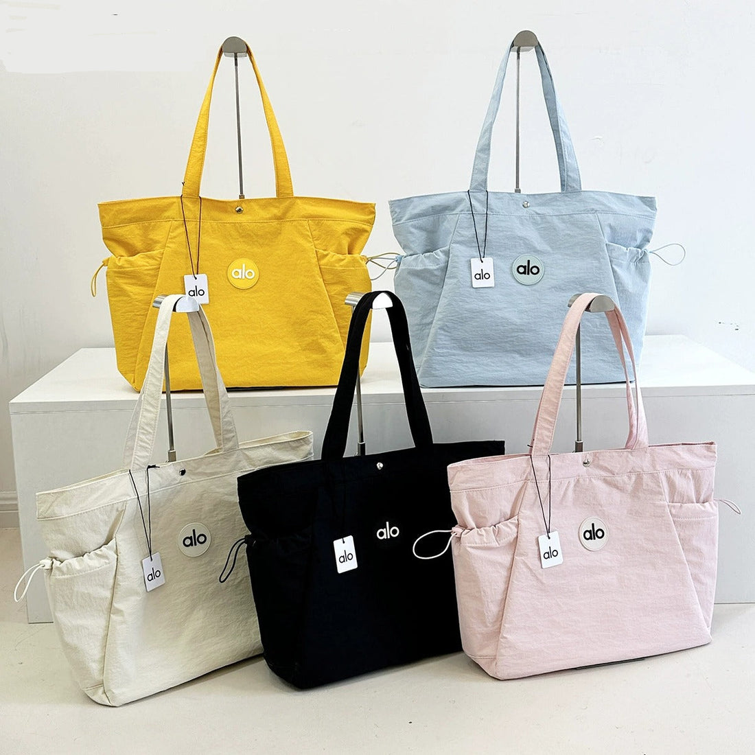 Five tote bags in yellow, blue, white, black, and pink on a white surface with a light gray background.