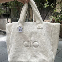 White fluffy tote bag with 'alo' branding held by a hand outdoors.
