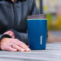 Blue insulated mug with brown lid on a table, held by a person wearing a black jacket.