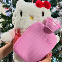 Hello Kitty plush toy holding a pink hot water bottle in front of a decorated Christmas tree.