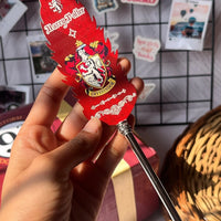 Hand holding a Harry Potter-themed pen with Gryffindor crest against a wall with photos and a basket.