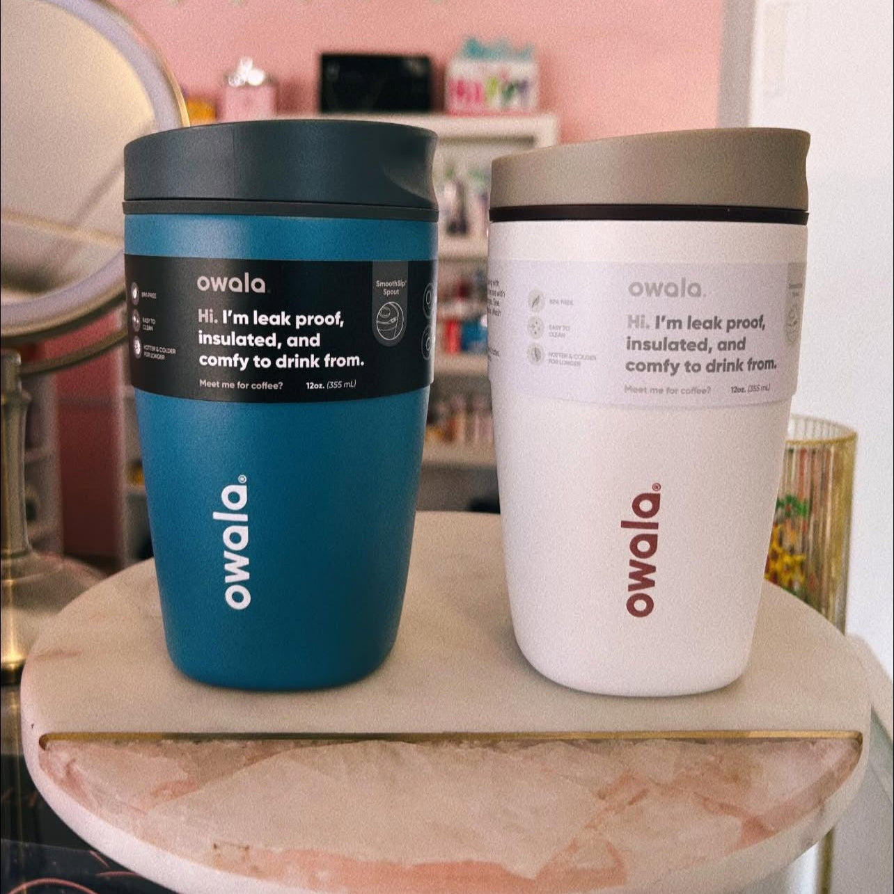 Two Owala tumblers, one blue and one white, on a marble surface with a blurred indoor background.