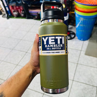 Green YETI Rambler bottle held by a person in an indoor setting with tiled floor and colorful buckets in the background.