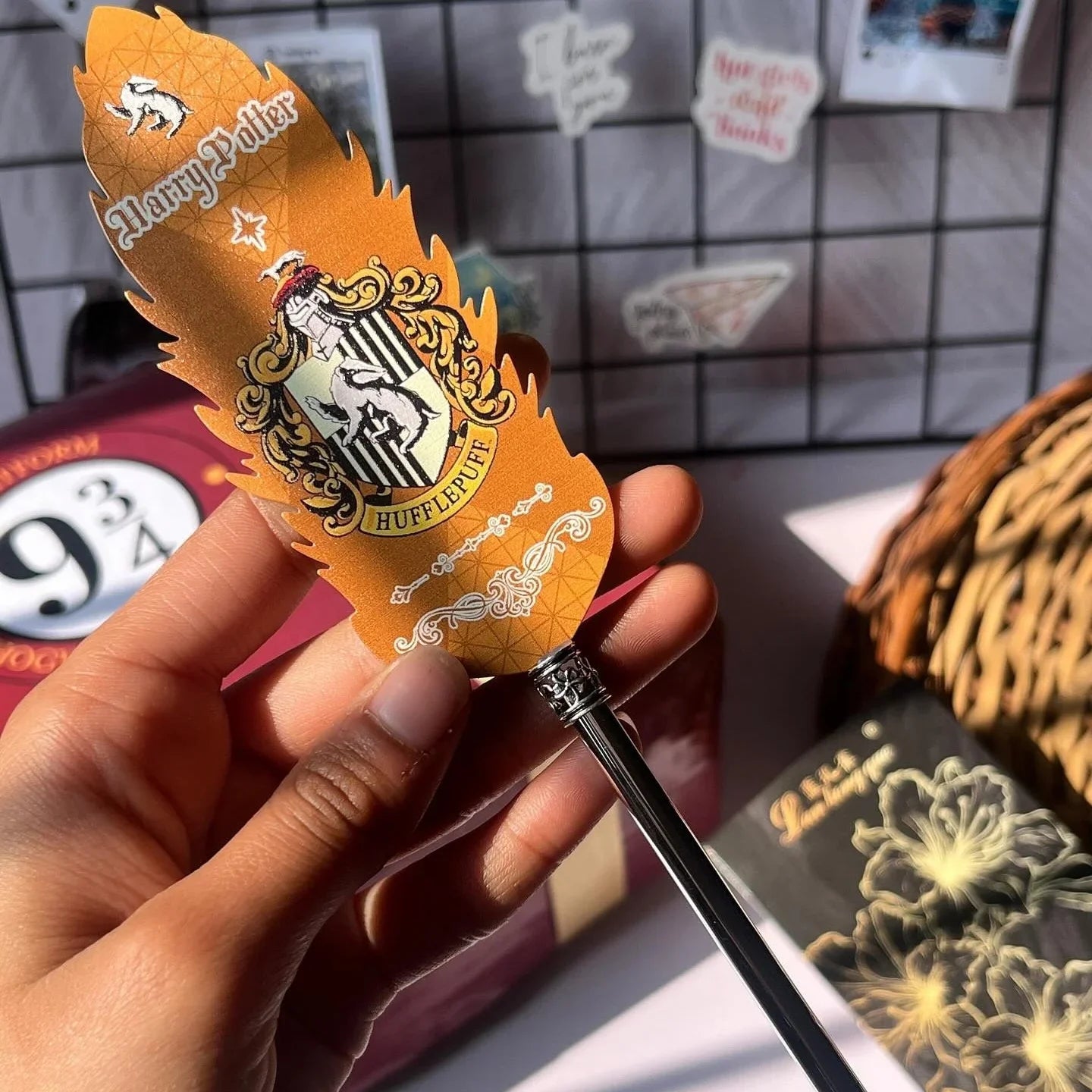 Hand holding a Harry Potter-themed quill with a decorative feather against a grid-patterned wall.