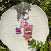 Pink bear keychain with purple heart charm on a white plate with green leaves in the background