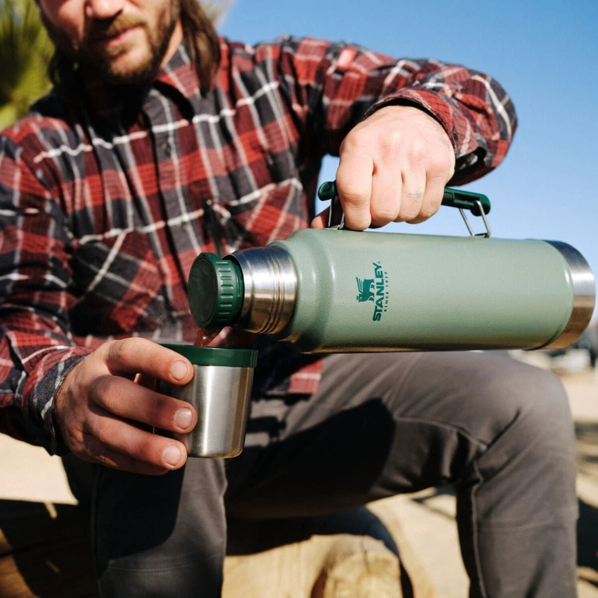 Person holding a green thermos outdoors with a clear sky background