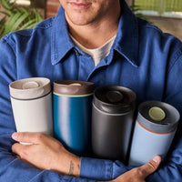 Person holding four different colored travel mugs outdoors.